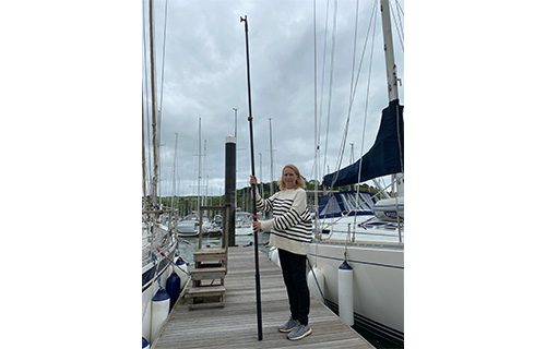 A person stands on a marina dock, holding a tall boat hook pole, surrounded by moored sailboats on a cloudy day.