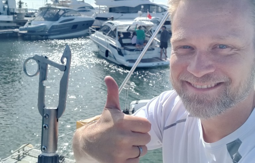 A man gives a thumbs up on a boat in the marina, surrounded by yachts. With a buoy catcher in hand, he navigates smoothly, mastering the waves and ensuring every knot is secure.