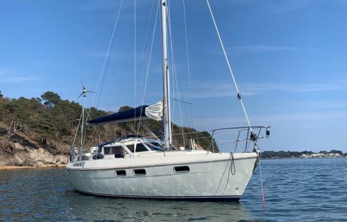 A white sailboat with blue accents is anchored in calm waters near a rocky shoreline under a clear blue sky, its boat mooring hook neatly secured.