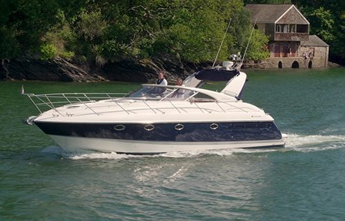 A sleek white motorboat with a black hull glides along the river, two people on board. A telescoping boat hook rests at the ready, useful for navigating tricky waters. In the background, a charming wooden boathouse nestles among the trees.