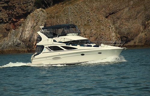 A sleek white motor yacht glides across the water, with rocky cliffs towering in the background, while a telescopic hook pole is ready on deck for seamless mooring.