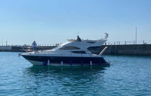 A motor yacht with two people on board is stationary on calm blue water near a concrete pier under a clear sky, effortlessly held in place by a boat mooring hook.