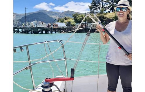 A woman wearing sunglasses and a hat stands on a boat, holding a telescopic hook pole. In the background, a marina and tree-covered hills stretch under a blue sky with clouds.