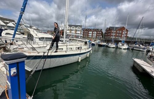 Person stands on a docked sailboat in a marina with several other boats, skillfully handling a boat hook pole. Buildings rise in the background under a partly cloudy sky.