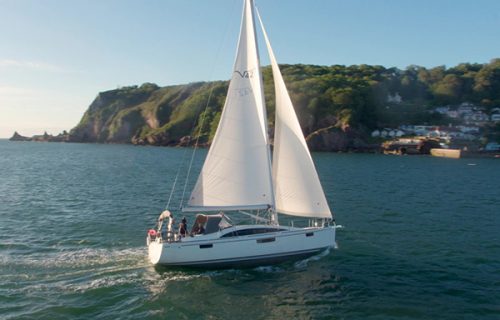 A white sailboat with two sails is gracefully navigating calm waters near a rocky coastline under a clear sky, expertly using its mooring buoy hook to make the journey even smoother.